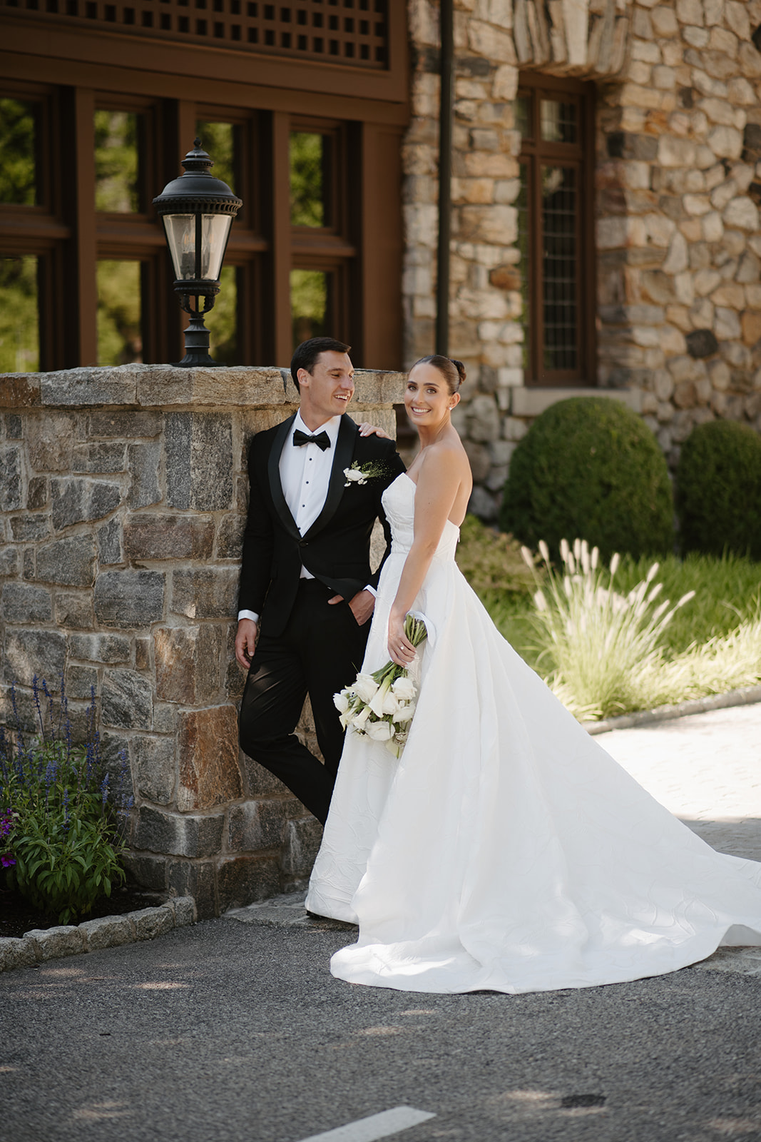 Groom looking lovingly at his smiling bride as they pose by a stone wall outside Le Chateau.